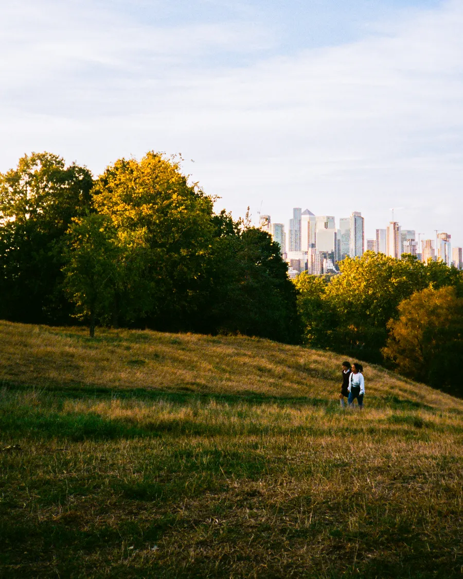 two people walking down a hill in greenwich park, london skyline beyond